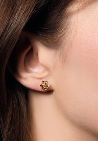 Złota kolczyk w kształcie węzła prezentowany na uchu, charakteryzujący się wielopętlowym wzorem o polerowanej powierzchni, podkreślającym jego gładką teksturę.
