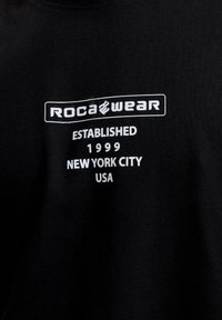 Mustapaitainen puuvillapaita, jossa on valkoista painettua tekstiä. Teksti sisältää "ROCA WEAR," "PERUSTETTU 1999," "NEW YORK CITY," ja "USA."