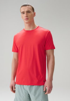 Ung mand med kort hår iført en ensfarvet rød t-shirt med korte ærmer og lysegrå shorts, stående foran en ensfarvet baggrund.