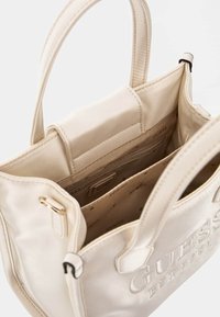 Borsa Guess di colore crema con doppio manico, logo stampato sul davanti e interni aperti che mostrano la fodera in tessuto e una tasca interna con zip.