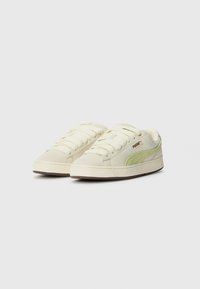 Cream-colored sneakers with a textured green suede accent, ribbed rubber sole, and flat cotton laces, featuring gold logo detailing.