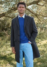 Lång, mörkgrå kappa med skräddarsydd design, bärs över en blå tröja, vit skjorta och ljusblå jeans, mot en naturlig bakgrund.