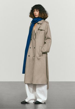 Femme aux cheveux bouclés portant un long manteau beige, une écharpe bleue, un pantalon large blanc et des chaussures noires pointues, debout devant un fond uni.