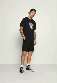 Zwarte katoenen t-shirt met een kleurrafische opdruk op de voorkant, gecombineerd met zwarte shorts en hoge zwarte sneakers met witte accenten.