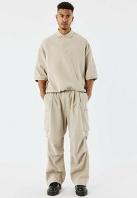Polo beige à manches courtes avec une texture côtelée, associé à un pantalon cargo ample assorti avec des poches latérales et une coupe décontractée. Chaussures noires.
