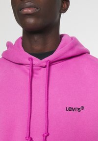Sweat à capuche rose avec cordons réglables et logo Levi's noir sur la poitrine. Comprend un tissu doux et une coupe standard.