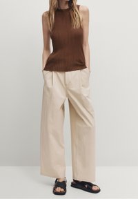 Kobieta w beżowych spodniach szerokich nogawkach z kieszeniami oraz brązowym bezrękawniku w prążki, w czarnych sandałach na krzyż, na jednolitym tle.