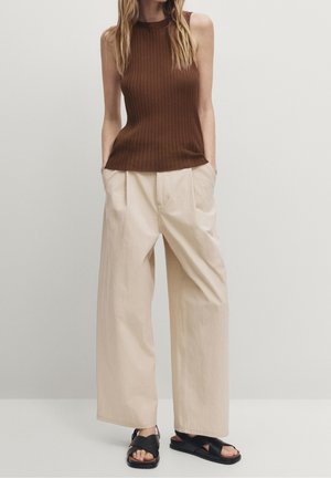 Woman wearing sleeveless brown ribbed top, beige wide-leg pants with pockets, and black cross-strap sandals against plain background.