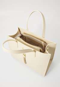 Beige synthetic tote bag with two handles, large main compartment, interior zippered pocket, and a gold tassel accent.