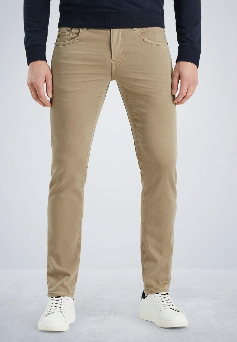 Pantalones de algodón color beige en corte ajustado, con bolsillos delanteros y una textura suave, combinados con zapatillas deportivas blancas, de pie frente a un fondo claro.