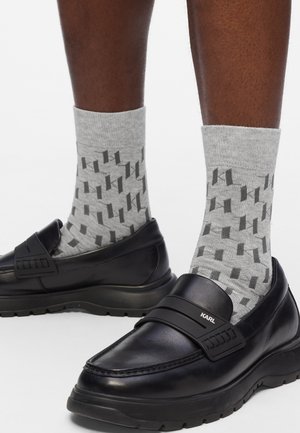 Mocassini in pelle nera con suola spessa e una fascia con il logo "KARL", indossati con calzini grigi che presentano un motivo geometrico scuro.