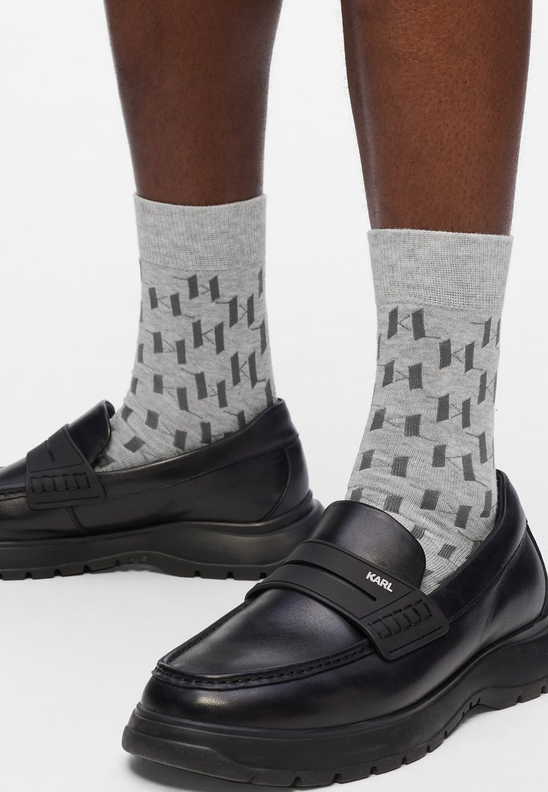 Mustad mustad nahast loafers paksu tallaga ja rihmaga, millel on "KARL" logo, kantud hallide sokkidega, millel on geomeetriline tume muster.