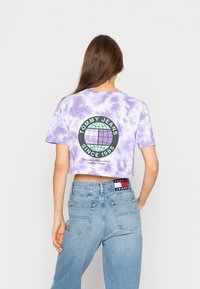 Fialové tričko s krátkým rukávem ve stylu tie-dye s černým kulatým logem, na kterém je zeměkoule a text. Spárováno se světle modrými džíny s vysokým pasem.