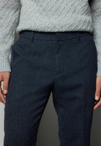 Marineblå uldblandede bukser med en skræddersyet pasform, der har en glat tekstur og rene linjer. Sammen med en lysegrå kabelstrikket sweater.