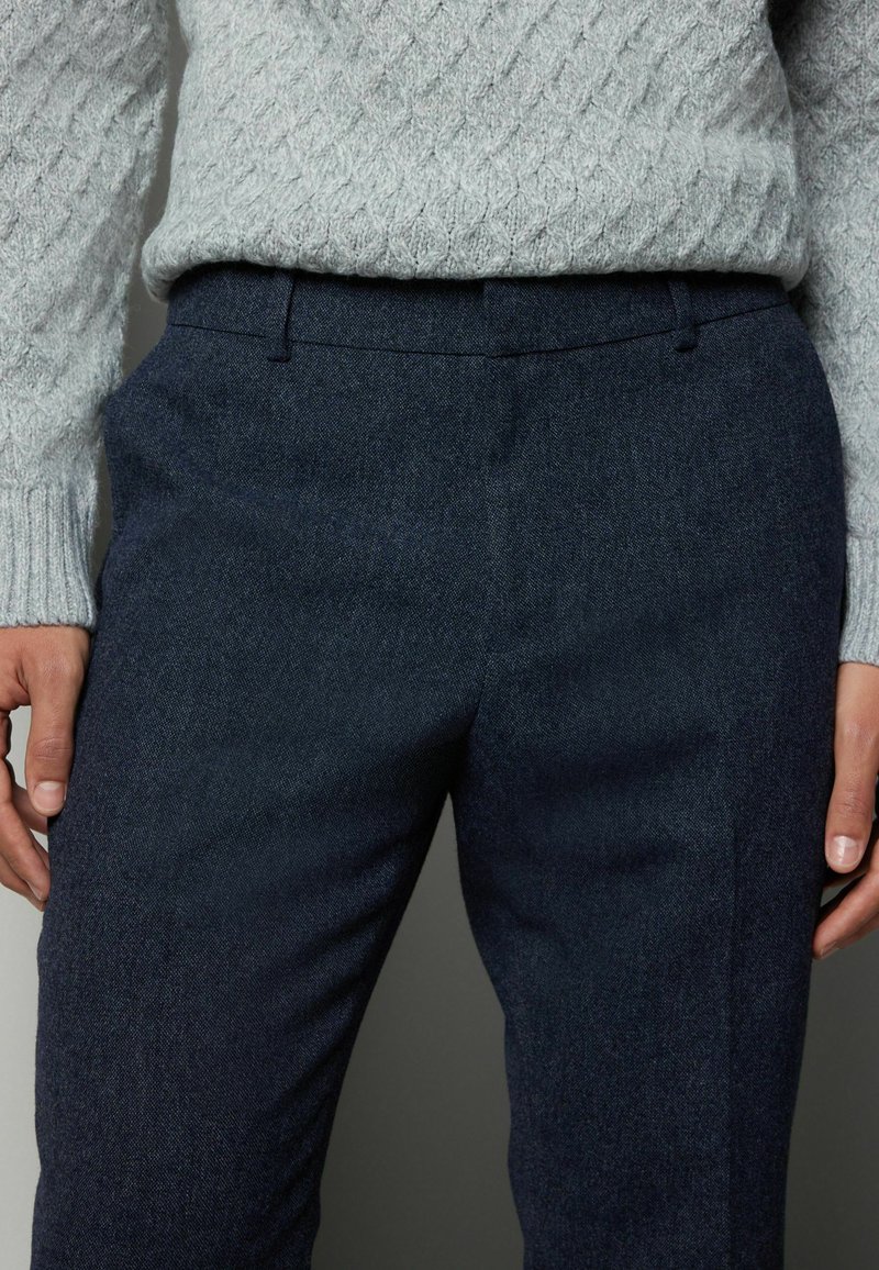 Marineblå uldblandede bukser med en skræddersyet pasform, der har en glat tekstur og rene linjer. Sammen med en lysegrå kabelstrikket sweater.