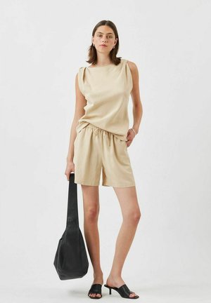 Beige sleeveless top with shoulder detailing and elastic waist shorts. Model holding a black shoulder bag, wearing black heeled mules.