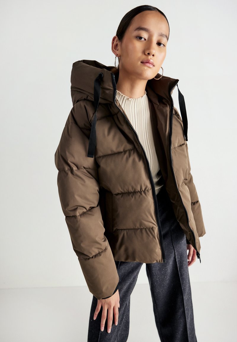 Brun pufferjakke med hætte, lynlåslukning og sorte snore. Har en quilted design og er parret med en ribbet cremefarvet top.