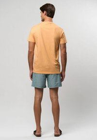 Vīrieša skats no mugurpuses, ģērbies gaiši oranžā kokvilnas T-kreklā un teļu svītrainās šortos. T-krekls ir ar vienkāršu dizainu un īsām piedurknēm.