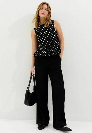 Femme portant un haut sans manches noir à pois, pantalon noir à jambes larges, tenant un sac à main noir, debout devant un mur blanc uni.