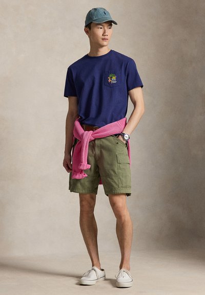 Mann med blå caps, marineblå T-skjorte med lomme-logo, olivengrønne cargoshorts, hvite sneakers og rosa genser knyttet rundt livet, står mot ensfarget bakgrunn.