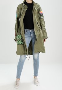 Osoba nosząca za dużą oliwkowozieloną kurtkę z łatami, czarną bluzkę, jasnoniebieskie dżinsy i białe sneakersy na jednolitym tle.