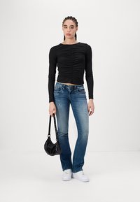 Black long-sleeve top with ruched front, paired with blue mid-rise jeans and white sneakers. Small black handbag with a shoulder strap.