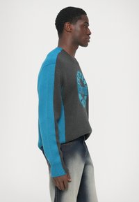 Junger Mann in blauem und grauem Langarmpullover mit kreisförmigem Logo, trägt ausgebleichte blaue Jeans und steht im Profil vor einem weißen Hintergrund.
