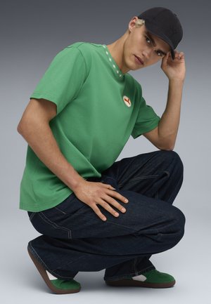 Camicetta verde a maniche corte con dettagli bianchi a forma di fiore, abbinata a jeans larghi blu scuro e scarpe verde e bianche. Cappellino nero indossato di lato.