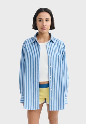 Frau steht nach vorne blickend und trägt ein blau-weiß vertikal gestreiftes Hemd über einem weißen bauchfreien Top und gelben Shorts.