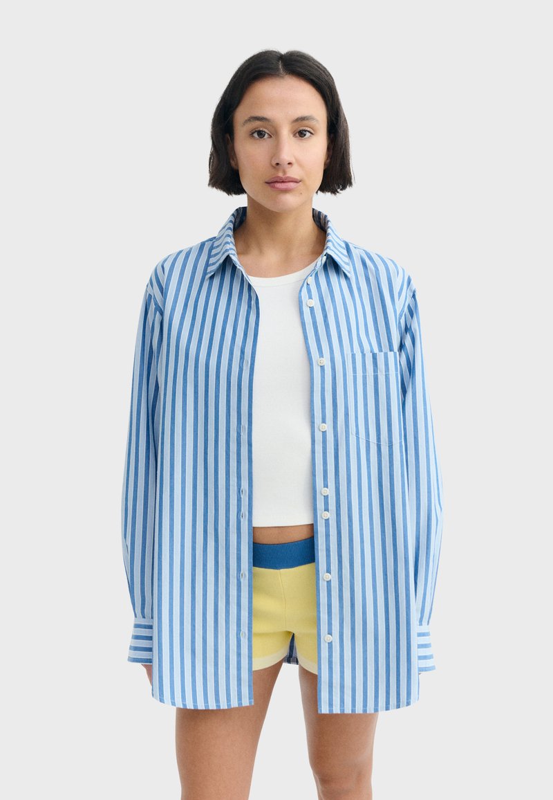 Femme debout, face à l'avant, portant une chemise rayée verticale bleue et blanche boutonnée sur un crop top blanc et un short jaune.