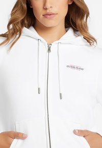 Balta zip-up džemperis, pagamintas iš minkštos medžiagos, su sidabriniu užtrauktuku ir raišteliais. Krūtinėje yra mažas raudonas ir juodas logotipas.
