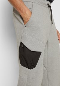Šedé tepláky s černou nylonovou kapsou, pasem na šnůrku a strukturovanou tkaninou. Viditelné boční kapsy a detaily šití.