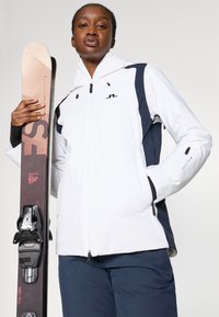 Balta slidinėjimo striukė su mėlynais akcentais, užtrauktukas priekyje ir reguliuojama kapišonu. Laiko rudos slidės su juodais tvirtinimais. Lygi tekstūra ir funkcinis dizainas.