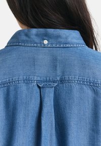Denim shirt with a light blue color, classic collar, single button at the neck, and a center pleat at the back for added detail.