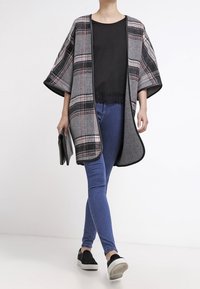 Šedý a kostkovaný plášť s volným střihem, spárovaný s černým topem a modrými skinny džíny. Černé slip-on boty dotvářejí outfit.