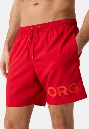 Homme portant un short de bain rouge vif avec une taille à cordon et "ORG" imprimé sur la jambe gauche, debout les mains dans les poches.