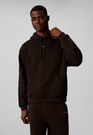 Man met een zwarte Calvin Klein hoodie en bijpassende broek, houdt de kraag van de hoodie vast, staand tegen een effen lichte achtergrond.