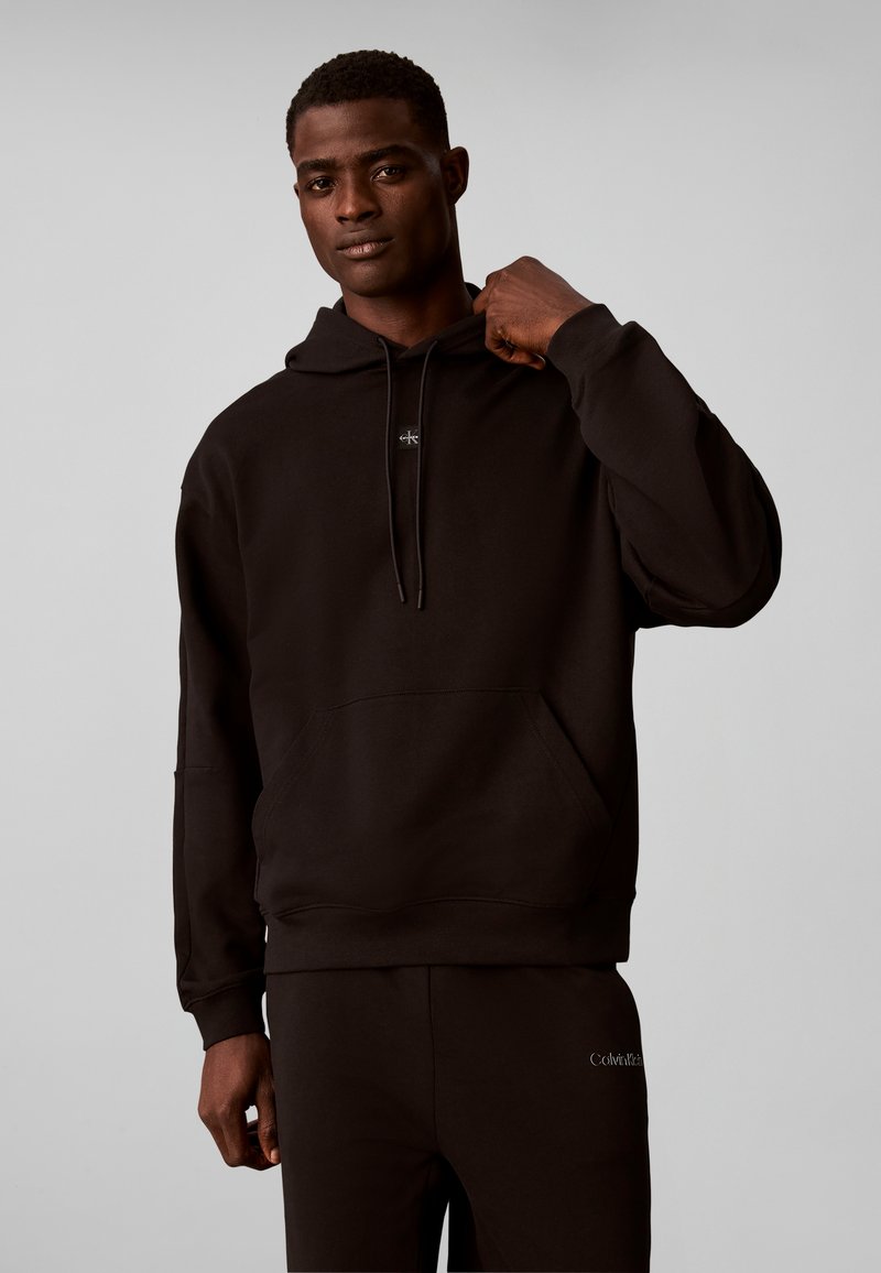 Homme portant un sweat à capuche noir Calvin Klein et un pantalon assorti, tenant le col du sweat à capuche, debout devant un fond clair uni.