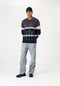 Pull en noir et gris tie-dye avec col rond ; jeans en denim bleu clair ; chaussures noires ; casquette noire simple ; texture lisse, coupe décontractée.