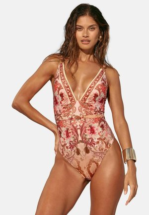 Kobieta z mokrymi włosami, ubrana w jednoczęściowy kostium kąpielowy z głębokim dekoltem w kształcie litery V, ozdobiony kwiatowym i paisley wzorem, z złotym paskiem i bransoletką na rękę.