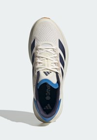 Chaussure de sport blanche avec des bandes bleu marine, des lacets beiges et un col de talon bleu vue de dessus sur un fond clair.