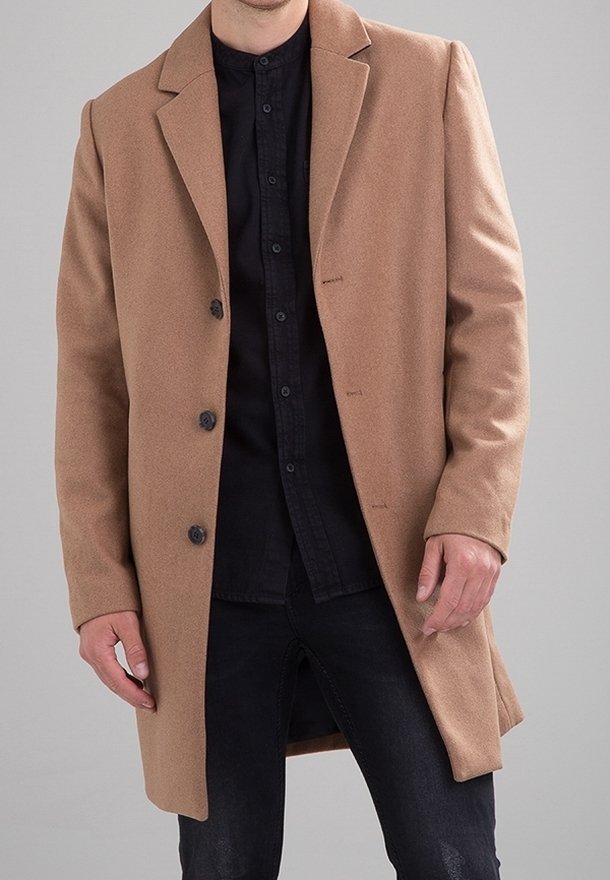 Homme portant un manteau beige mi-genou par-dessus une chemise noire boutonnée et un pantalon noir, sur un fond gris uni.