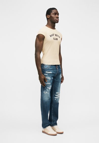 Ανδρικό μοντέλο που φοράει αμάνικο μπεζ τοπ με την επιγραφή «Bad Boys Club», φθαρμένο μπλε τζιν και μπεζ αθλητικά παπούτσια, στέκεται μπροστά σε απλό λευκό φόντο.