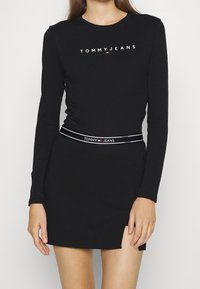 Musta pitkähihaine mekko, jossa on pyöreä kaula-aukko ja sivuaukot. Varustettu kontrastilla "TOMMY JEANS" -logolla ja raidallisella vyötärönauhalla.