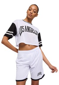 Top corto in rete bianca con maniche corte nere e "LOS ANGELES" in bold lettering nero. I pantaloni corti bianchi presentano un orlo nero e un testo abbinato.