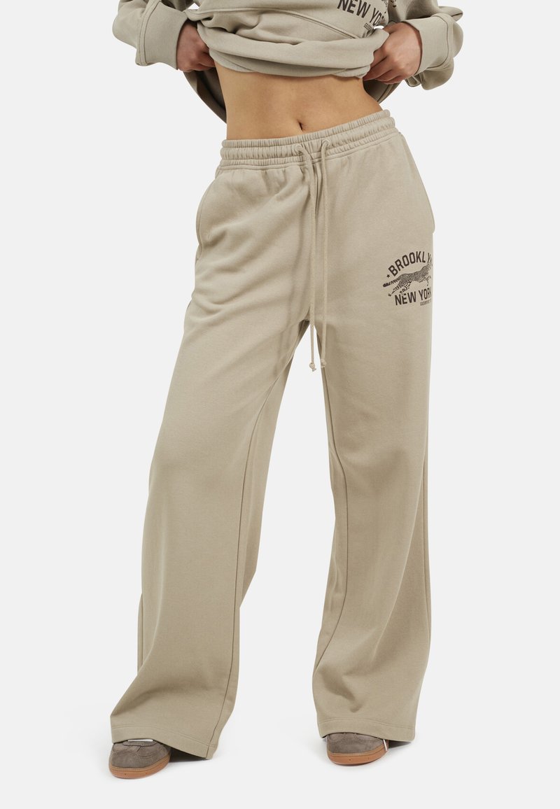 Beige wijde sweatpants met een drawstring tailleband en de tekst "Brooklyn New York" en een luipaardgrafiek op de linker dij, gedragen met grijze sneakers.
