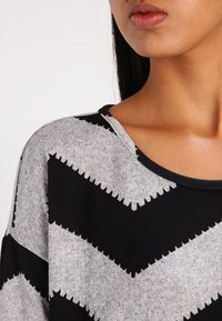 Nahaufnahme einer Person, die einen grauen Pullover mit schwarzen Zickzackmustern und einem weiten runden Ausschnitt trägt, wobei das untere Gesicht und der Hals sichtbar sind.