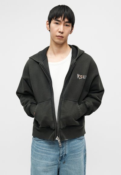 Σκούρο πράσινο φούτερ με φερμουάρ από απαλό ύφασμα, με μεγάλο κουκούλα, τσέπες στο μπροστινό μέρος και μικρό λογότυπο στο αριστερό στήθος. Στυλιζαρισμένο με ανοιχτόχρωμο τζιν.