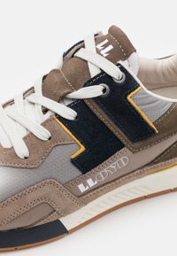 La sneaker présente une combinaison de daim marron, d'accents bleu marine, de tissu texturé gris, de lacets blancs et d'un logo distinctif sur le côté.