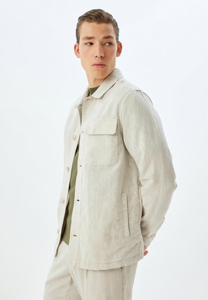 Giacca button-up beige chiaro in un tessuto testurizzato. Presenta una tasca sul petto e tasche laterali. Indossata sopra una camicia verde. Design sottile e casual.